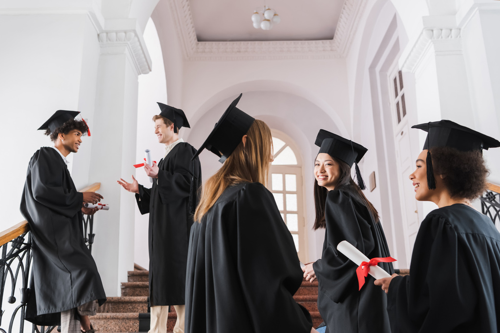 Міжрасові випускники в академічних костюмах і шапочках розмовляють в університеті
  - Фото, зображення