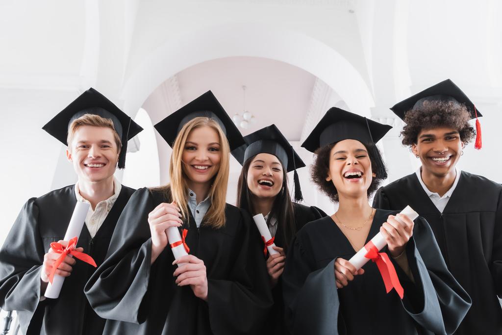 Estudiantes multiculturales positivos con diplomas sonriendo a la cámara  - Foto, Imagen