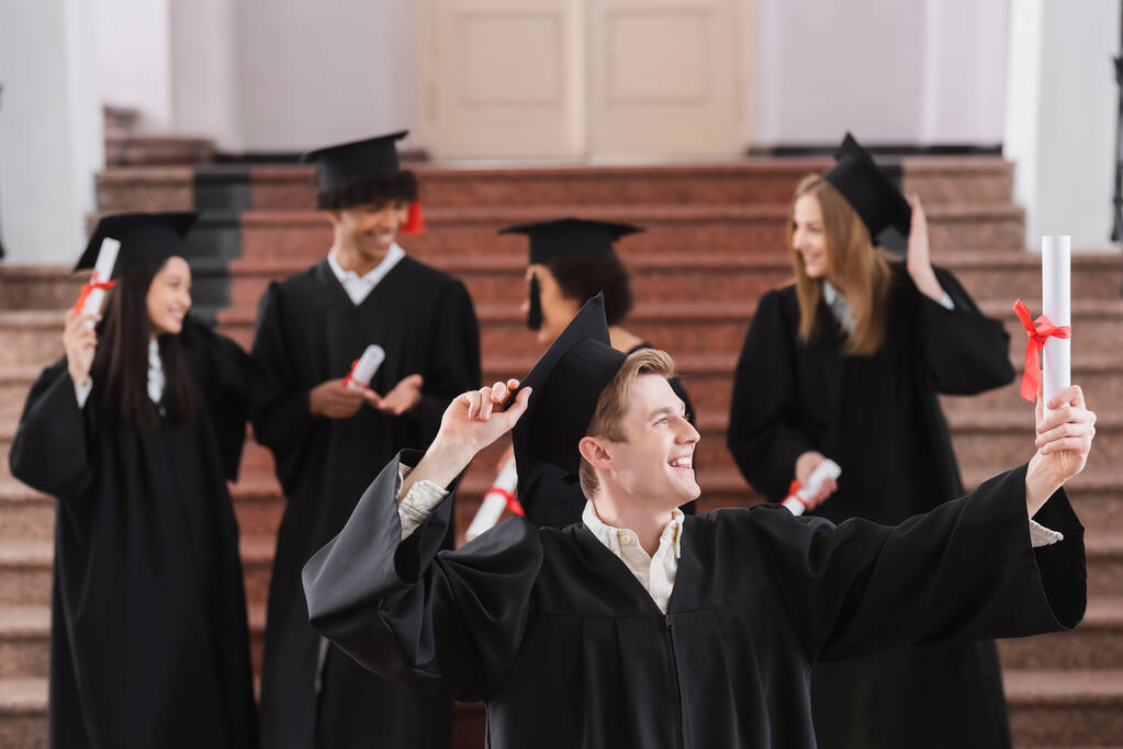 Bachelor lächelt, während er Diplom in der Nähe von Freunden auf verschwommenem Hintergrund betrachtet  - Foto, Bild