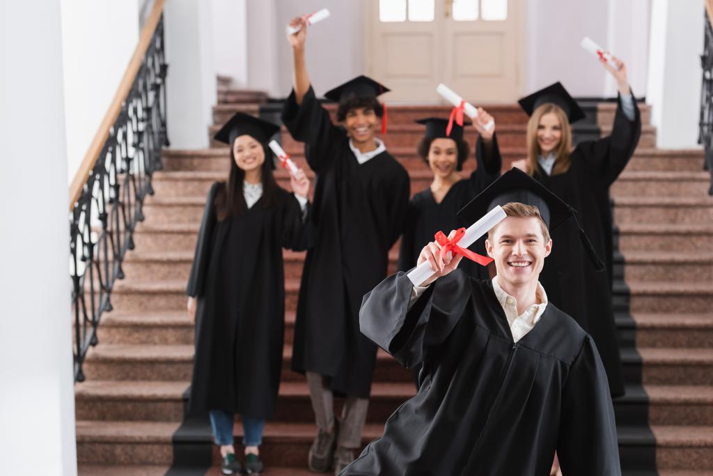 Licenciado con diploma sonriendo cerca de estudiantes interracial en un fondo borroso  - Foto, imagen