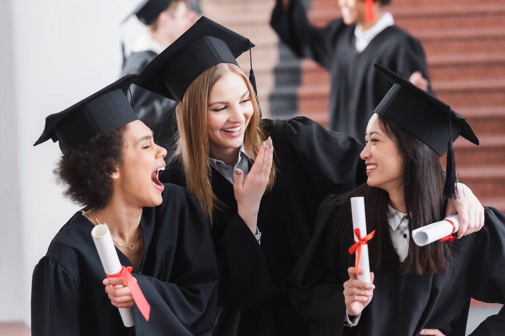 Возбужденные многонациональные выпускники с дипломами, смеющиеся в университетском зале - Фото, изображение