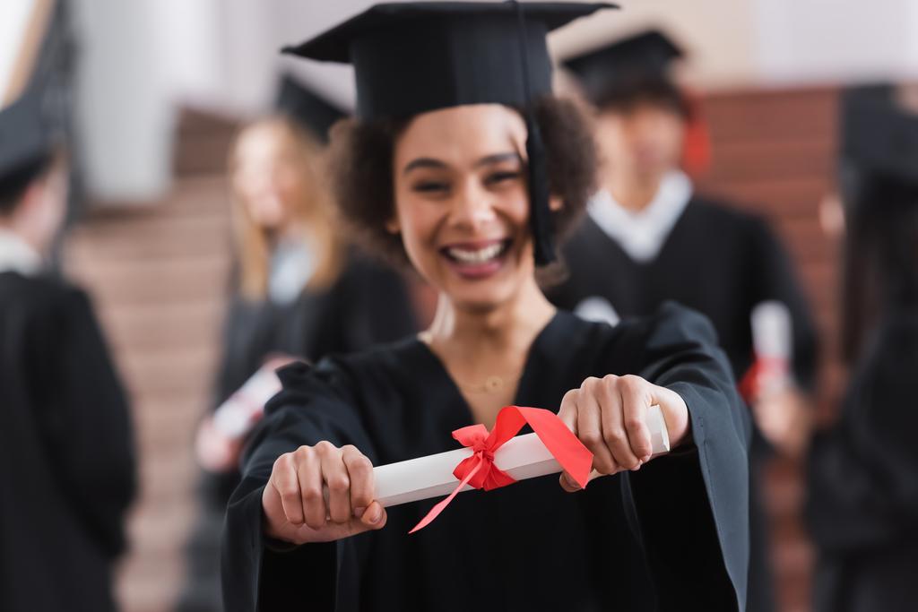 Diploma met lint in handen van vrolijke Afrikaans-Amerikaanse student op wazig achtergrond  - Foto, afbeelding