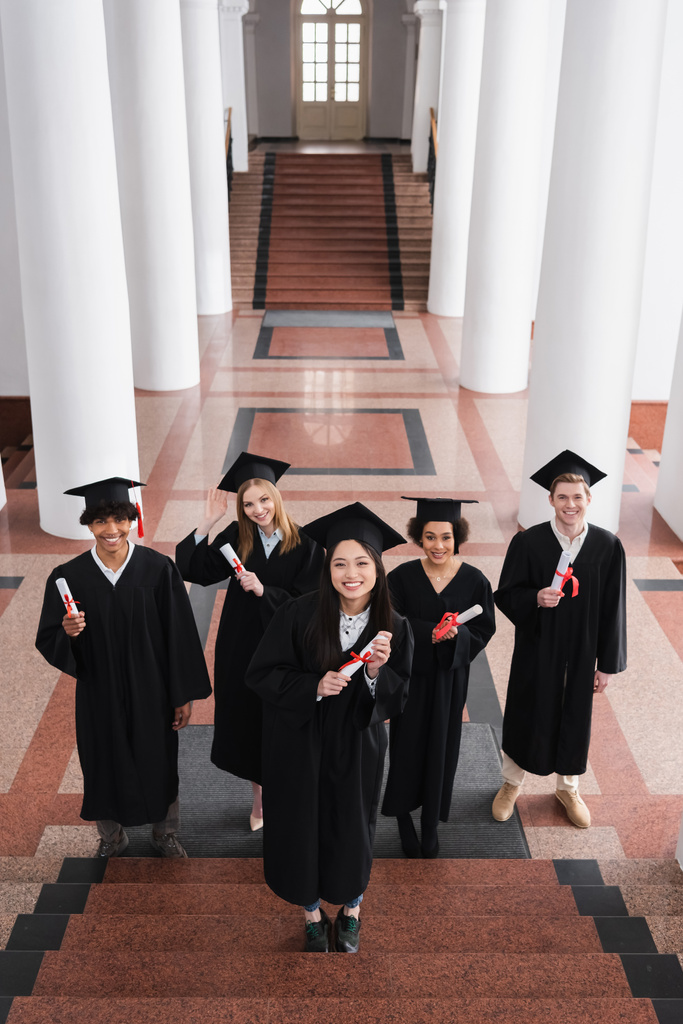 Vista ad alto angolo di scapoli multietnici sorridenti con diplomi sulle scale dell'università  - Foto, immagini