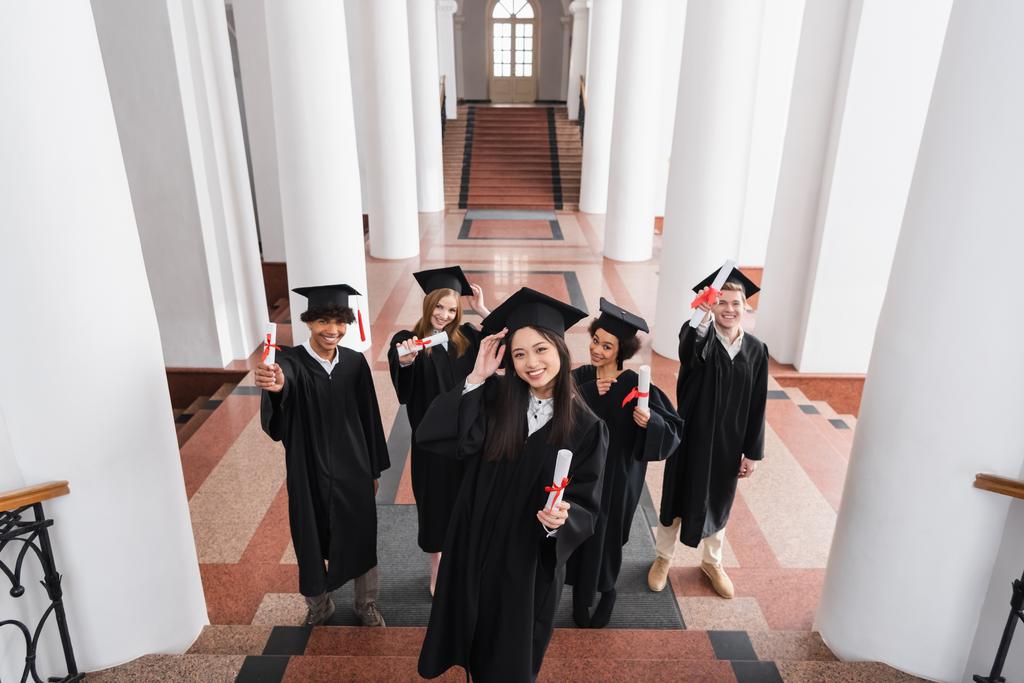 Blick aus der Vogelperspektive auf fröhliche asiatische Hochschulabsolventen mit Diplom in der Nähe von Freunden in Mützen  - Foto, Bild