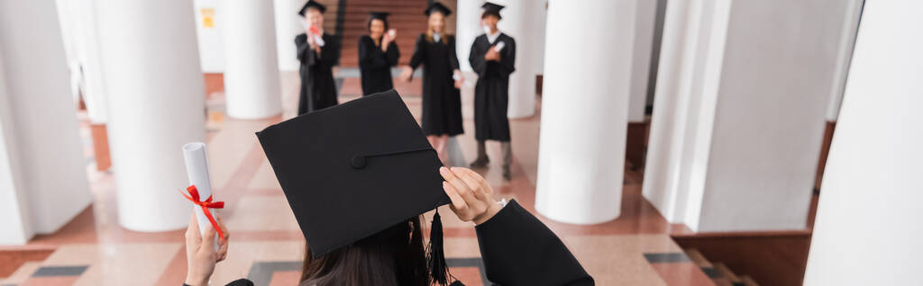 Achteraanzicht van student in diploma afstuderen cap holding diploma in de buurt van wazig vrienden, banner  - Foto, afbeelding