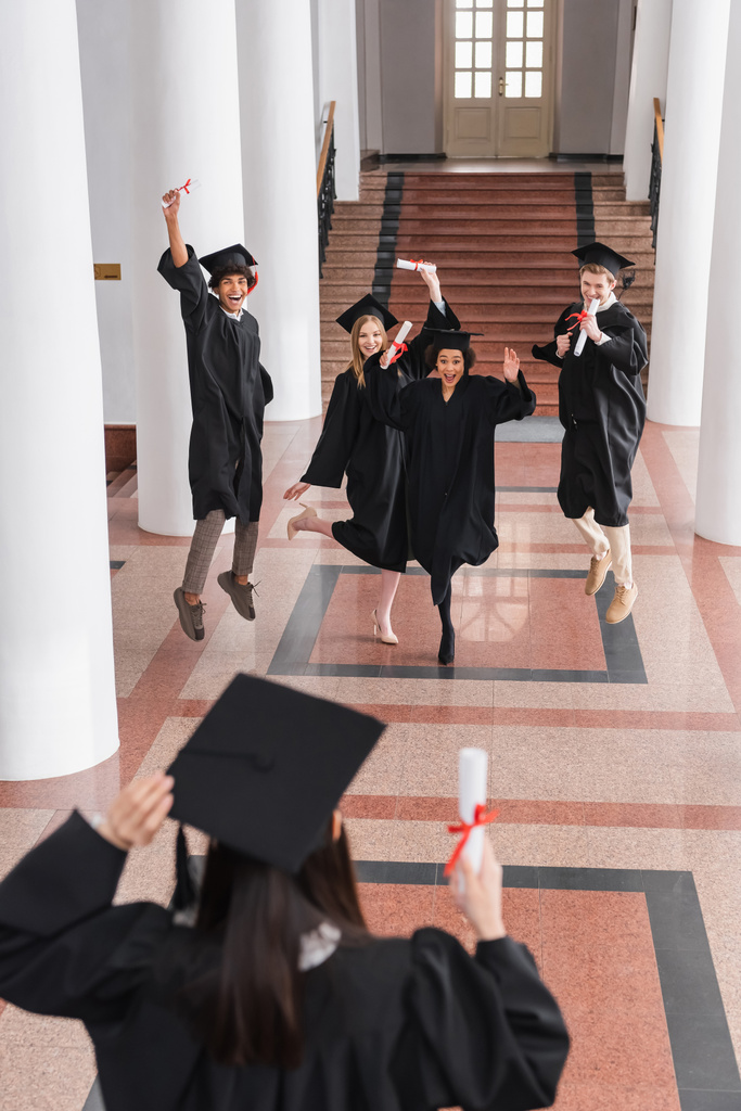 Formatos multiétnicos excitados com diplomas pulando perto de amigo em primeiro plano desfocado - Foto, Imagem
