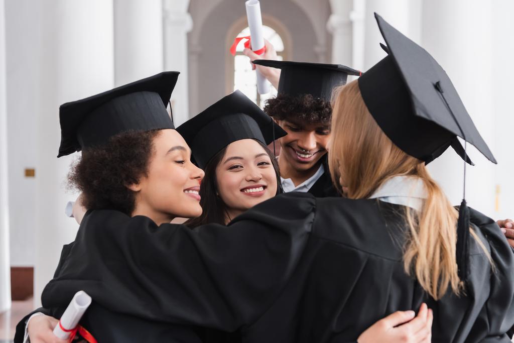 Cheerful asian graduate looking at camera while hugging friends  - Photo, Image