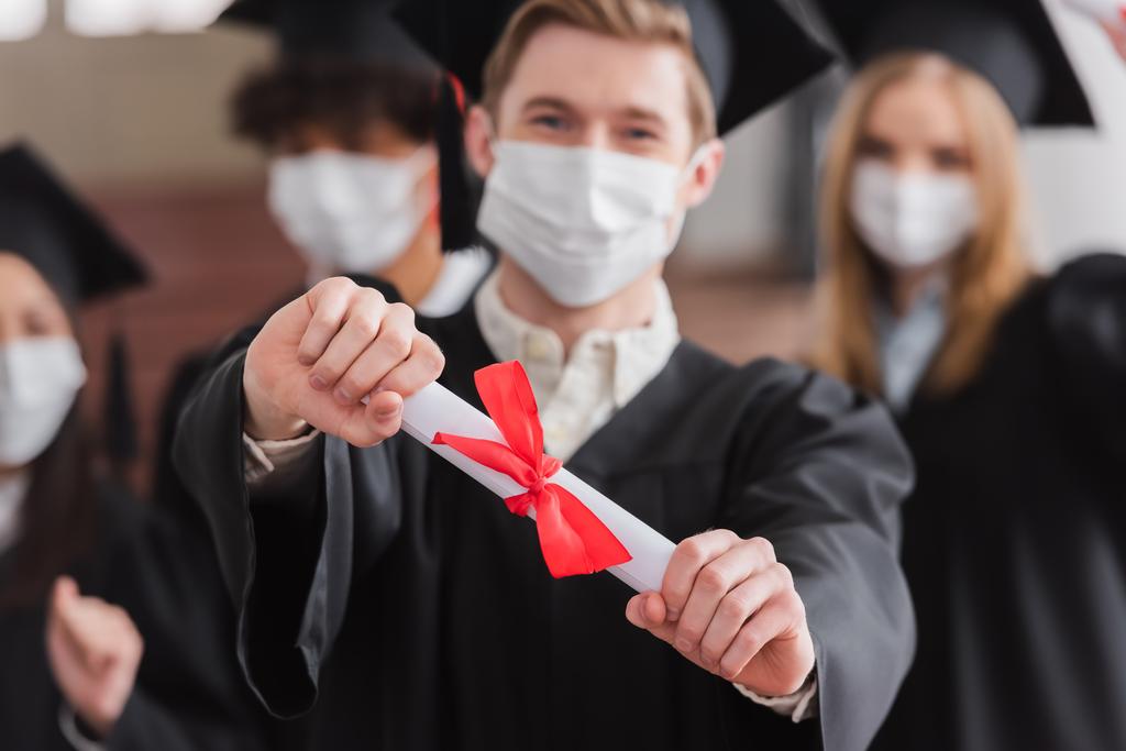 Diploma en mano de soltero en máscara médica sobre fondo borroso  - Foto, imagen