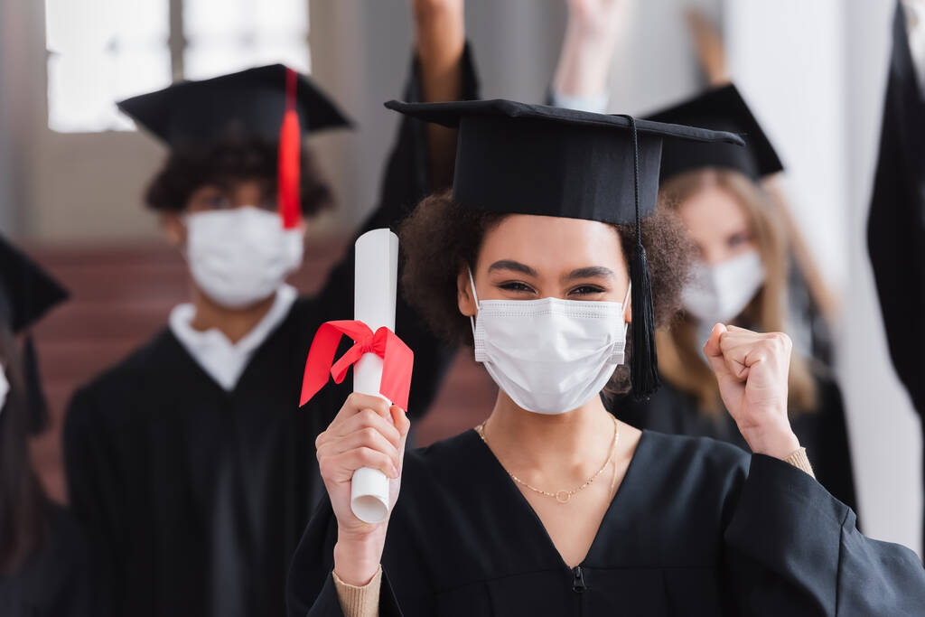 Graduado afroamericano en máscara médica con diploma y gesto de sí  - Foto, imagen