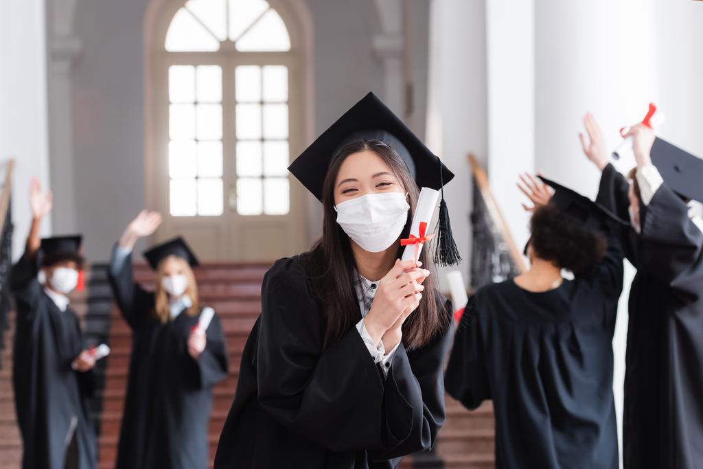 Aziatisch bachelor in cap en medisch masker houden diploma in de buurt wazig vrienden  - Foto, afbeelding