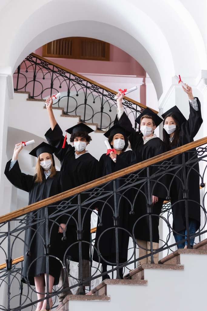 Multicultural students in medical masks and academic caps holding diplomas on stairs in university  - Photo, Image