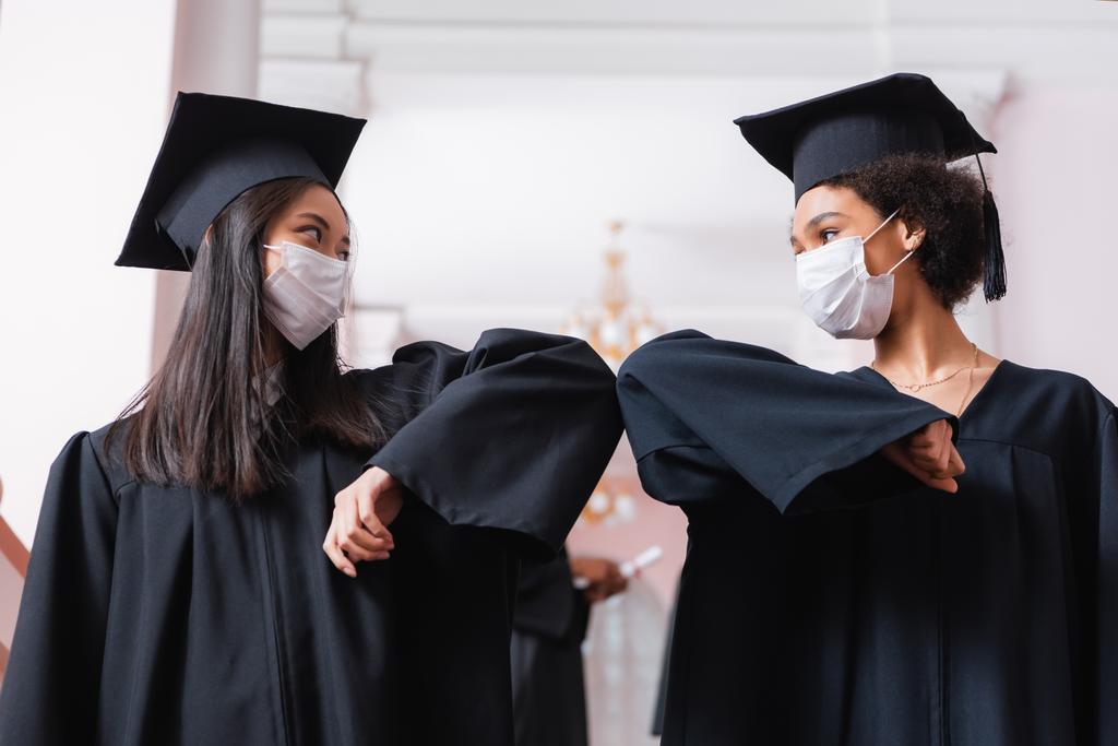 Afrikanische und asiatische Absolventen in medizinischen Masken beim Ellbogenschlag  - Foto, Bild
