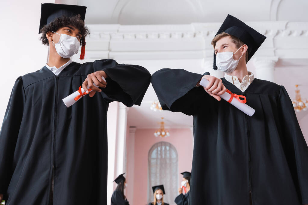 Vue à angle bas de célibataires interracial dans les masques médicaux faisant bosse du coude tout en tenant des diplômes  - Photo, image