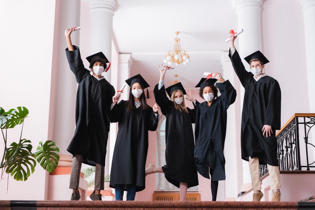 Wieloetniczni absolwenci w maskach medycznych podnoszący dyplomy na uniwersytecie  - Zdjęcie, obraz