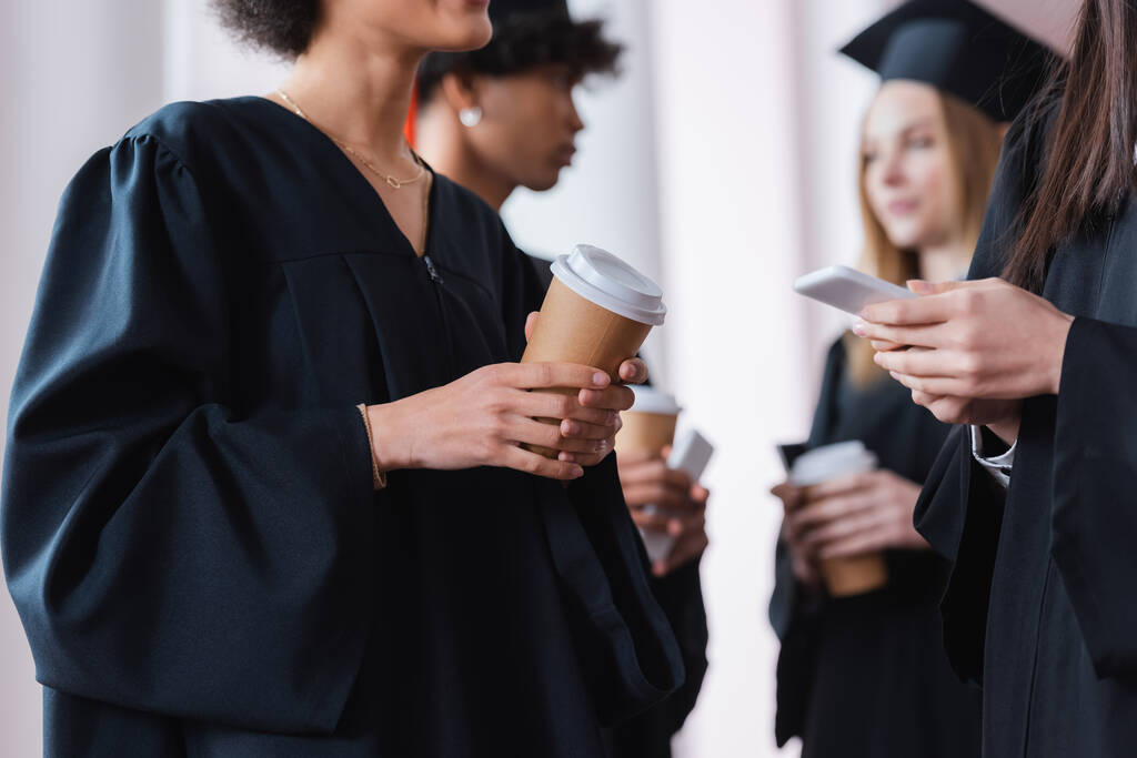 Мультиэтнические выпускники держа кофе, чтобы пойти и смартфон  - Фото, изображение