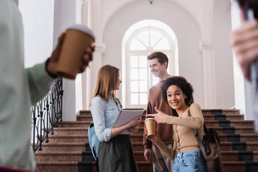 glimlachende Afrikaans-Amerikaanse student met koffie te gaan wijzen in de buurt van vrienden in de universiteit  - Foto, afbeelding