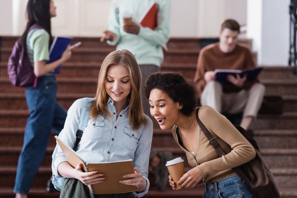 Excited african american student with coffee standing near friend with notebook  - Photo, Image