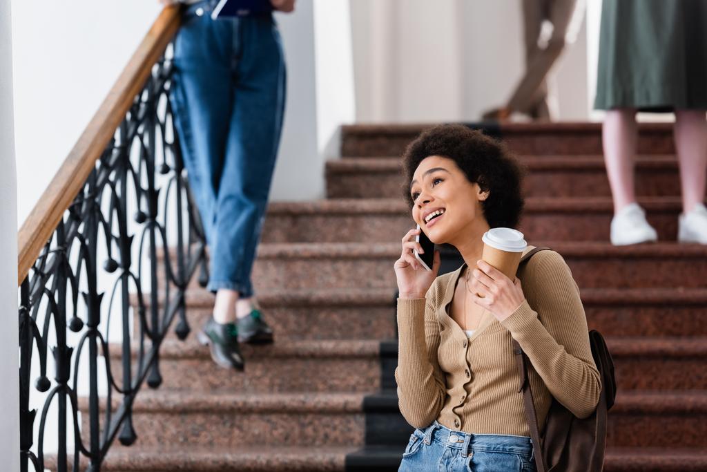Usmívající se africký americký student mluví na mobilu a drží kávu jít na univerzitu  - Fotografie, Obrázek