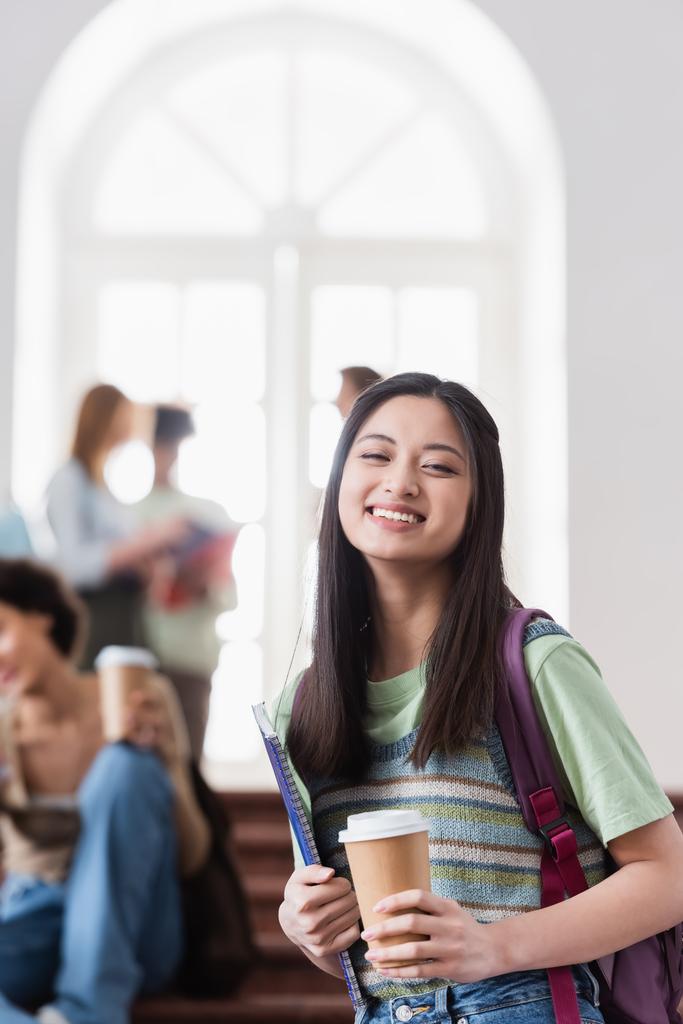 Sorridente studente asiatico con tazza di carta e notebook guardando la fotocamera  - Foto, immagini