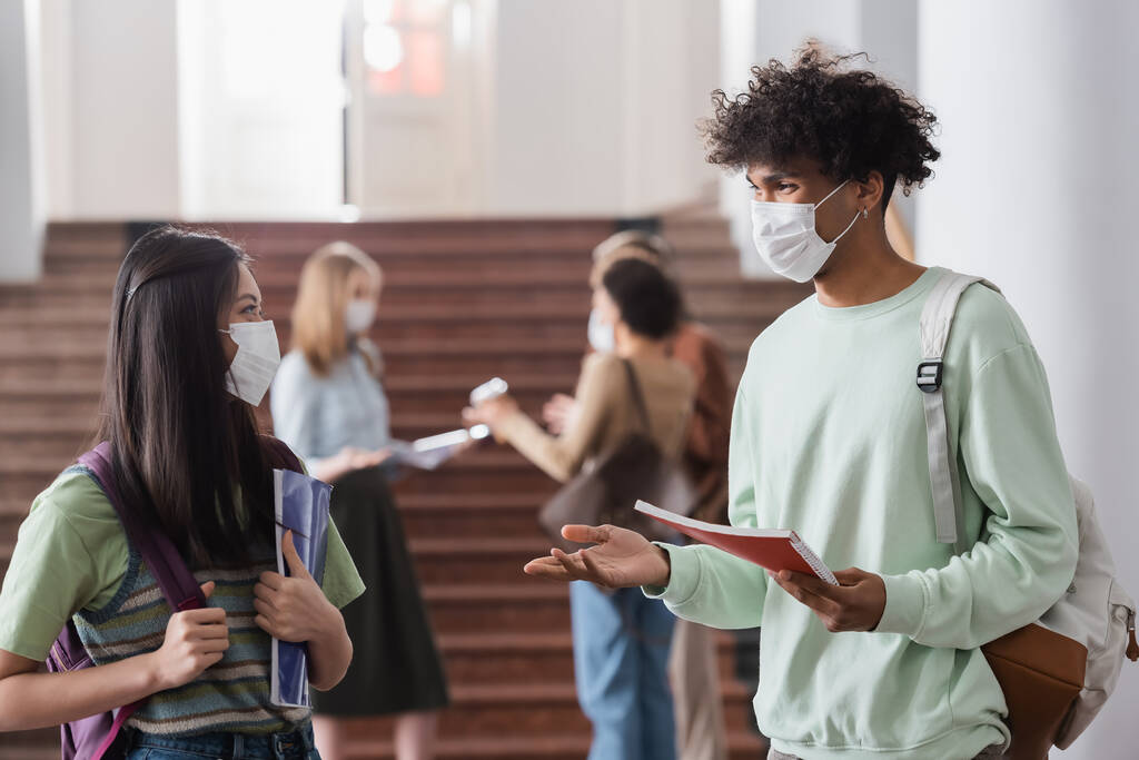 Πολυεθνικοί φοιτητές με ιατρικές μάσκες που κρατούν σημειωματάρια και μιλάνε  - Φωτογραφία, εικόνα