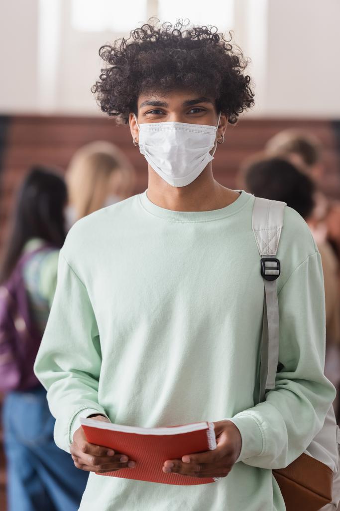 Studente afroamericano in maschera medica in possesso di notebook  - Foto, immagini