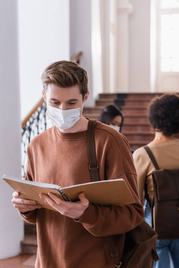 Φοιτητής με ιατρική μάσκα κοιτάζοντας σημειωματάριο κατά τη διάρκεια καραντίνας στο πανεπιστήμιο  - Φωτογραφία, εικόνα