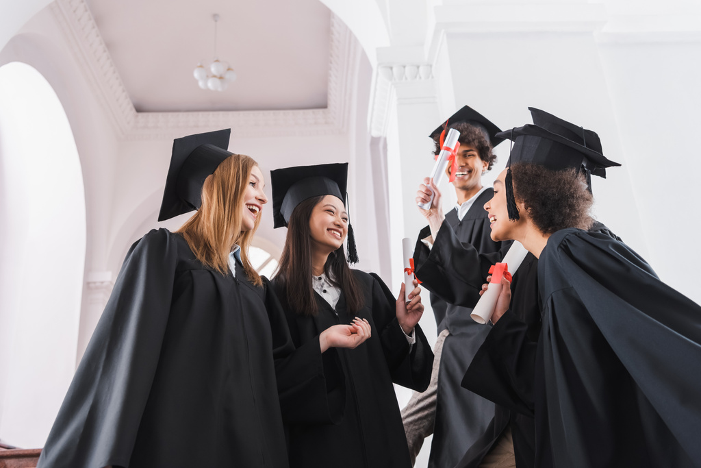 Niski kąt widzenia uśmiechniętych absolwentów międzyrasowych z dyplomami w hali uniwersyteckiej  - Zdjęcie, obraz