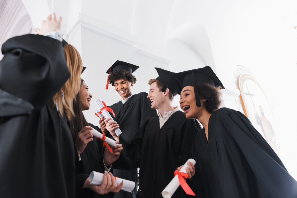 Vista de ángulo bajo de los graduados multiétnicos felices con diplomas en el salón de la universidad  - Foto, Imagen
