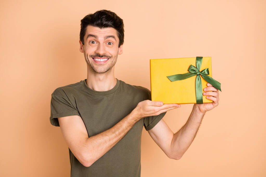 Photo de gars démontrer cadeau boîte toothy sourire regarder caméra porter vert t-shirt isolé beige couleur fond - Photo, image