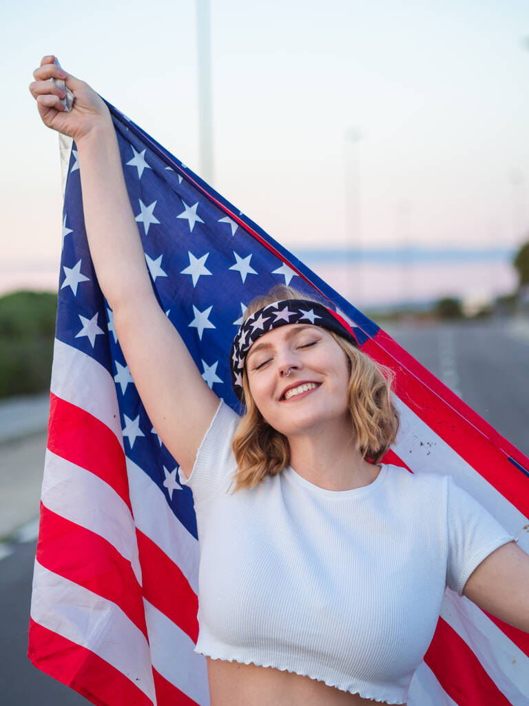 Μια κάθετη φωτογραφία μιας ξανθιάς Καυκάσιας από την Ισπανία να κρατάει την αμερικανική σημαία πίσω της και να χαμογελάει με τα μάτια κλειστά. - Φωτογραφία, εικόνα