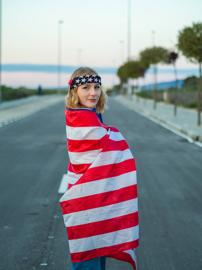 Un colpo verticale di una donna caucasica bionda spagnola coperta dalla bandiera americana mentre indossa un cerchietto USA - Foto, immagini