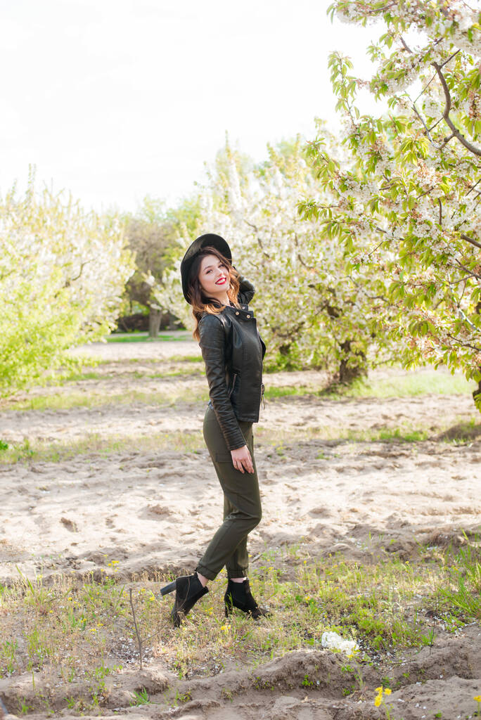 Милая молодая девушка в черной шляпе и модной черной кожаной куртке весной в цветущем вишневом саду в солнечную погоду с цветущей вишневой веткой в руках. Весна и мода - Фото, изображение