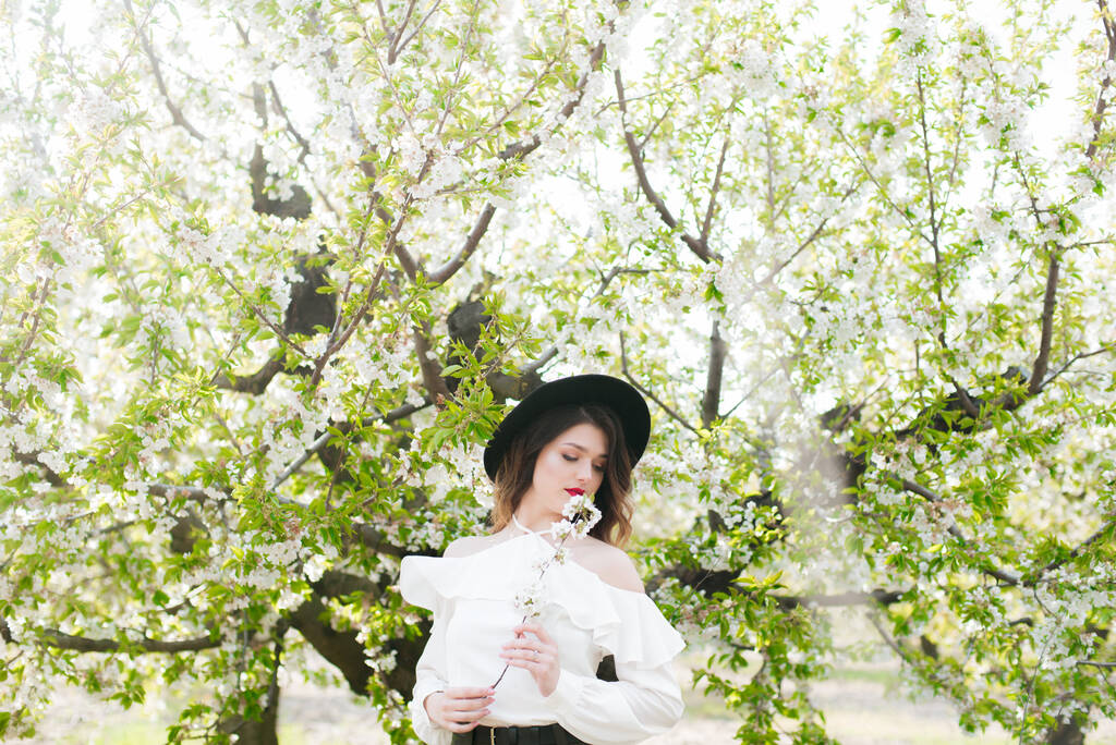 Gyönyörű fiatal lány fekete kalapban és fehér, romantikus selyemblúzban tavasszal egy virágzó cseresznyekertben, napsütéses időben, virágzó cseresznyeággal a kezében. Tavasz és divat - Fotó, kép