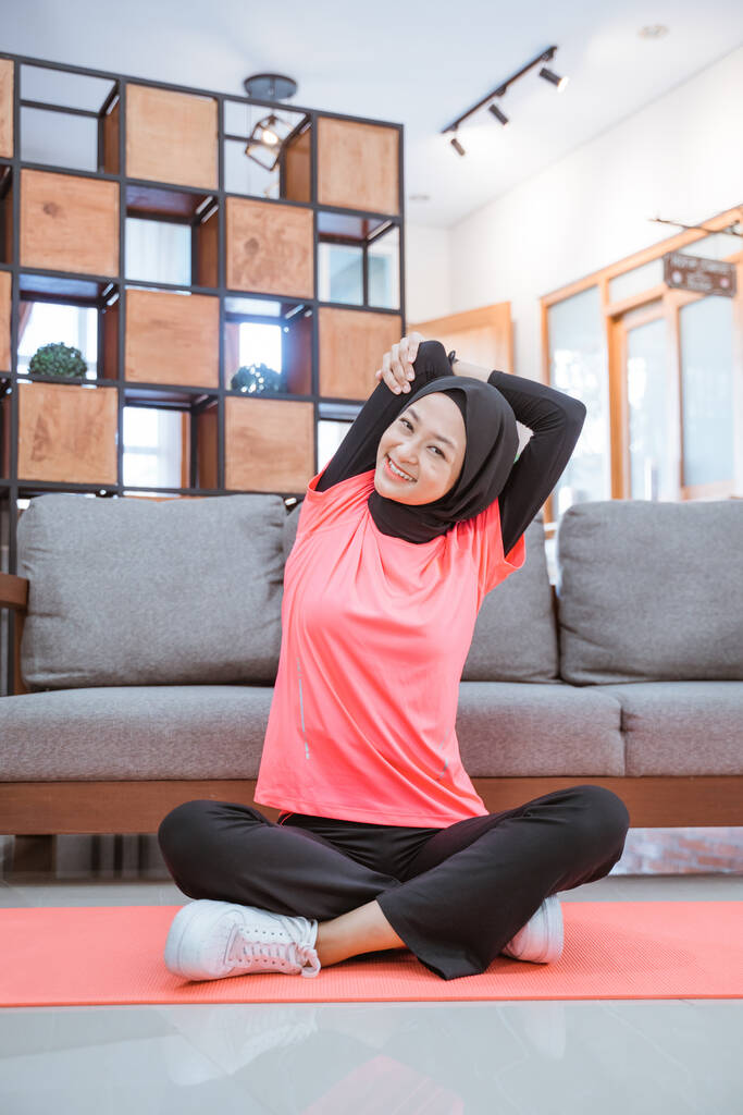 ein Mädchen im verschleierten Fitnessstudio-Outfit lächelt beim Dehnen der Hand, während es mit einer Matte auf dem Boden sitzt - Foto, Bild