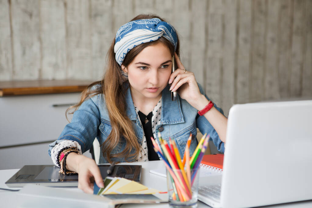Портрет молодої красивої щасливої усміхненої випадкової жінки дизайнер сидить за столом з інструментами, кольоровими зразками і ноутбуком
 - Фото, зображення