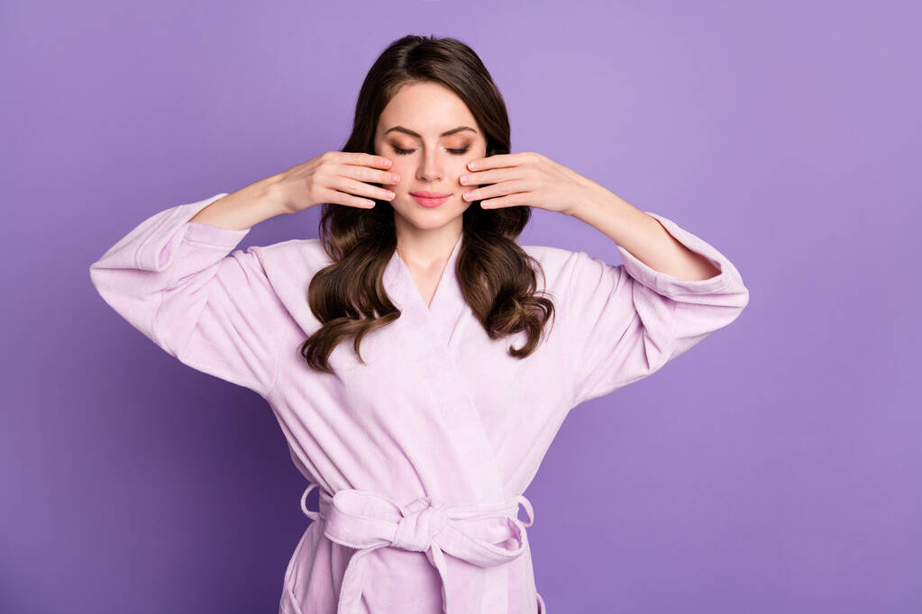 Portrait photo de femme les yeux fermés appliquant de la lotion sur les joues avec deux mains isolées sur fond violet vif - Photo, image