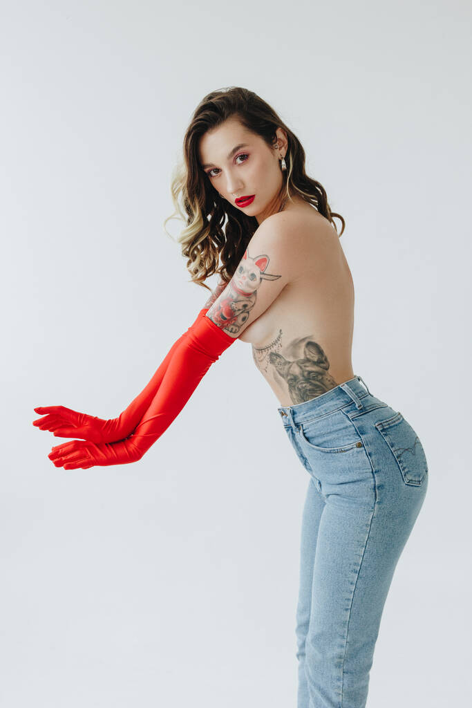 Sexy žena s tetováním na sobě džíny a červené rukavice na bílém pozadí - Fotografie, Obrázek