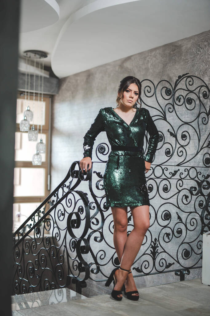 An adult woman wearing a sparkling green dress with high heels and leaning on floral handrails in a restaurant - Photo, Image