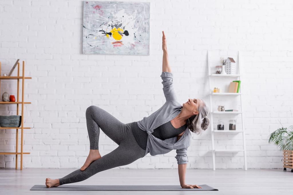 flessibile donna di mezza età in abbigliamento sportivo con mano sollevata praticare yoga sul tappeto a casa  - Foto, immagini
