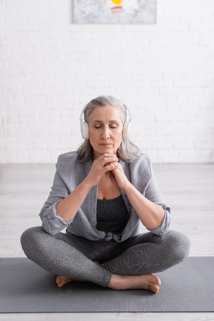 Frau mittleren Alters meditiert mit drahtlosen Kopfhörern, während sie in Lotuspose auf einer Yogamatte sitzt  - Foto, Bild