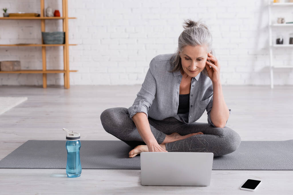 Frau mittleren Alters in Sportbekleidung sitzt in Lotus-Pose auf Yogamatte und schaut Online-Tutorial auf Laptop  - Foto, Bild