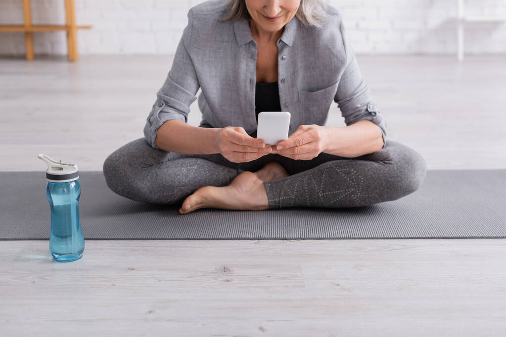 abgeschnittene Ansicht einer Frau mittleren Alters, die in Lotuspose auf einer Yogamatte sitzt und ihr Smartphone benutzt  - Foto, Bild