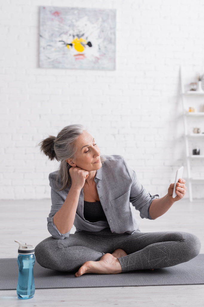 middle aged woman sitting in lotus pose on yoga mat and using smartphone  - Photo, Image