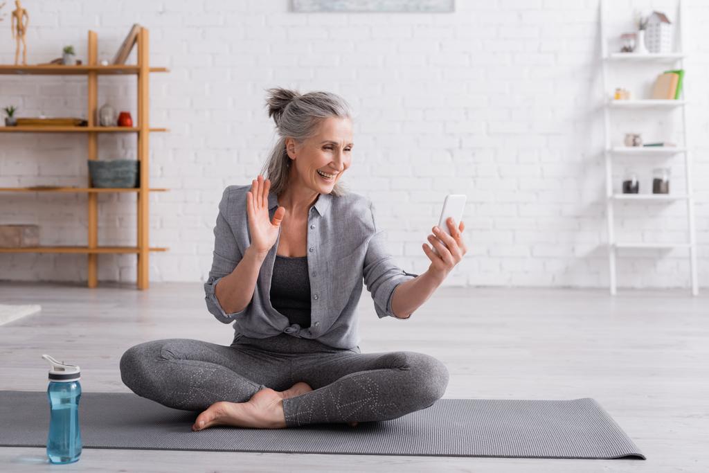 mujer de mediana edad sentada en una pose de loto en una esterilla de yoga y saludando con la mano mientras recibe una videollamada en un teléfono inteligente  - Foto, Imagen