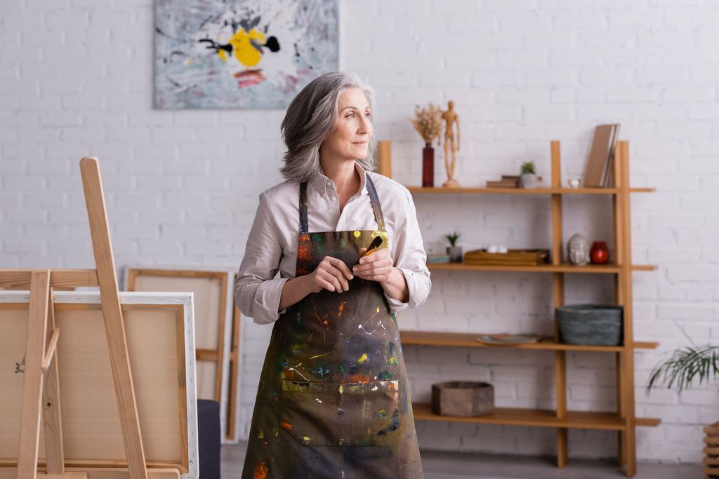 middle aged artist in apron holding paintbrush near easel  - Photo, Image