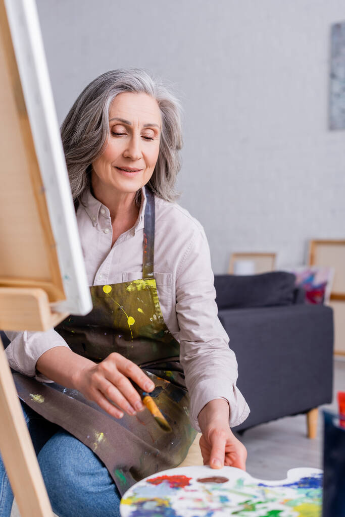 Künstler mittleren Alters hält Pinsel und reicht Palette mit bunten Farben  - Foto, Bild