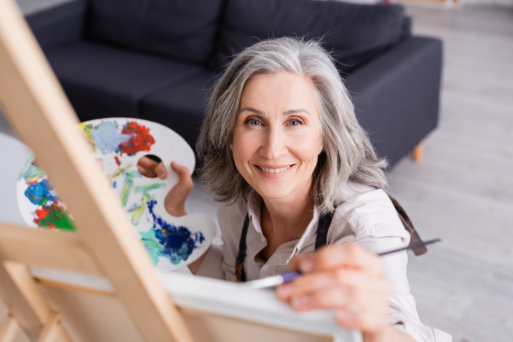 χαμογελαστή ώριμη γυναίκα που κρατάει πινέλο και παλέτα ενώ ζωγραφίζει με θολή πρόσοψη  - Φωτογραφία, εικόνα