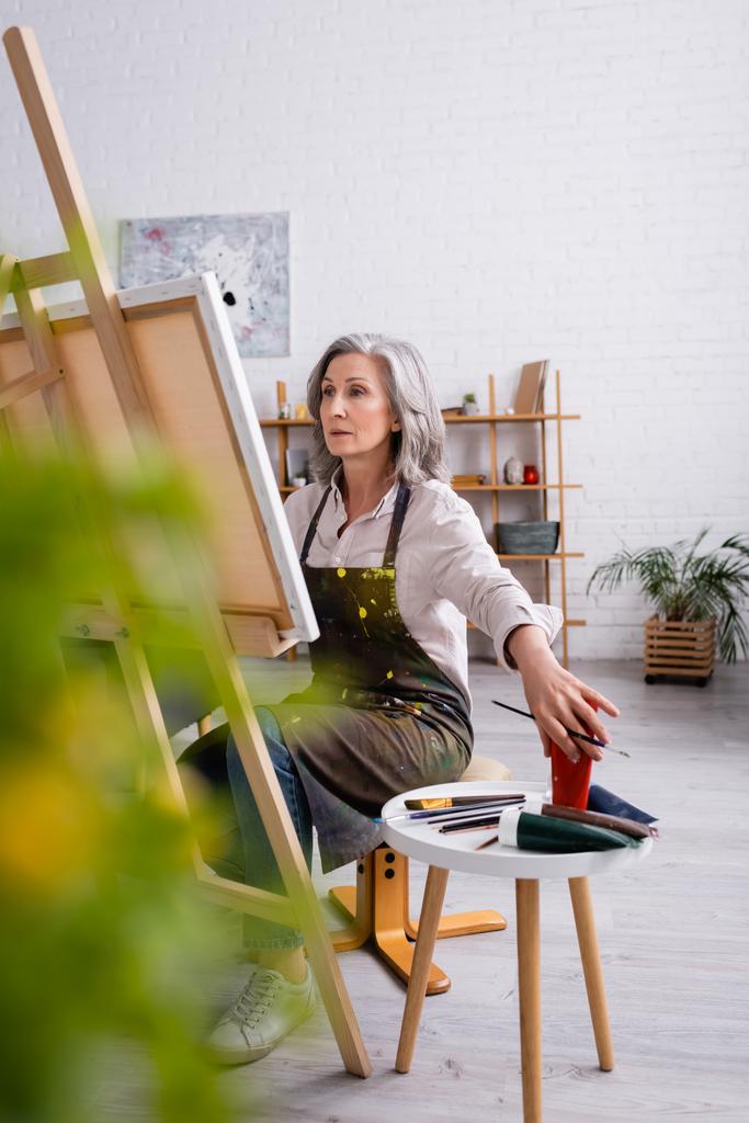 artista maduro sosteniendo pincel y alcanzando la pintura en tubo mientras está sentado cerca de la lona  - Foto, imagen
