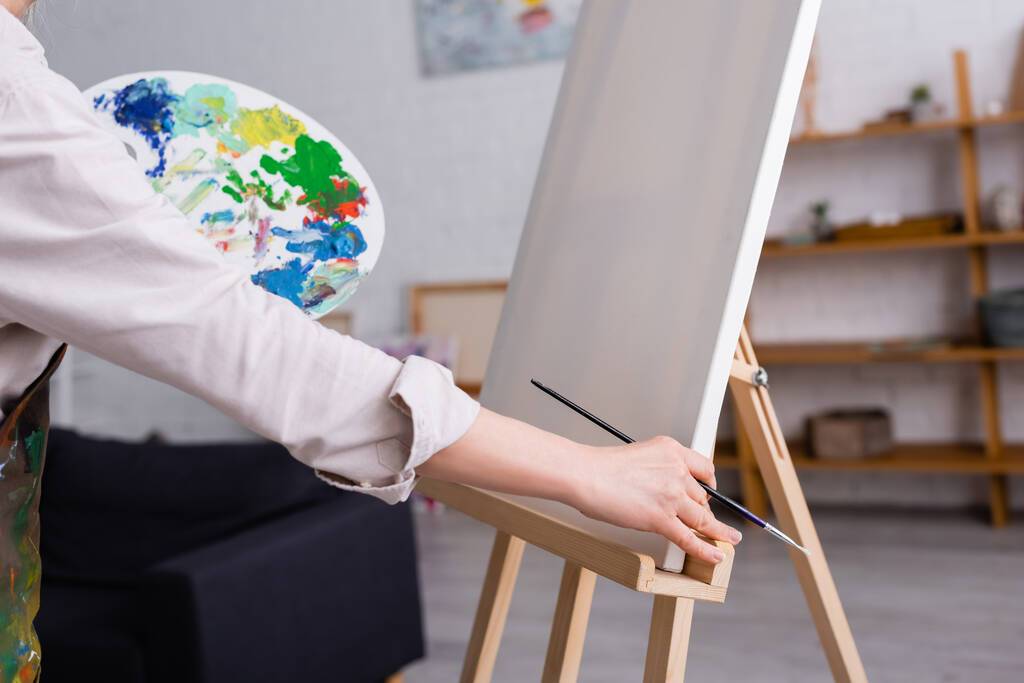 gedeeltelijk zicht op vrouw van middelbare leeftijd met penseel en palet met kleurrijke verf in de buurt van blank doek  - Foto, afbeelding