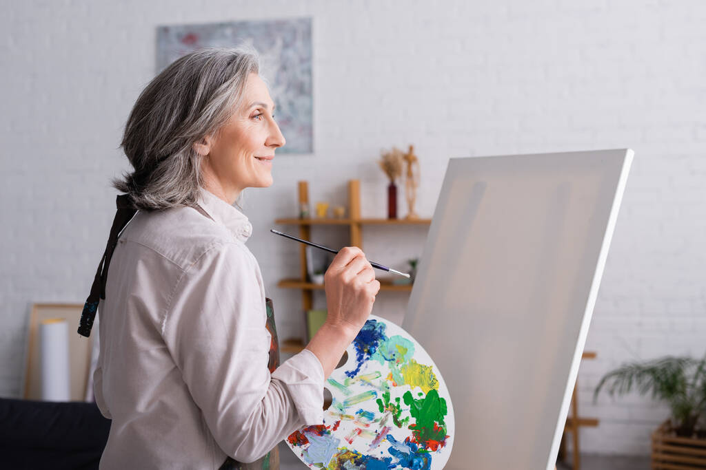Orta yaşlı bir kadın elinde boya fırçası ve palet ile boş tuvalin yanında renkli boyalar tutuyor.  - Fotoğraf, Görsel